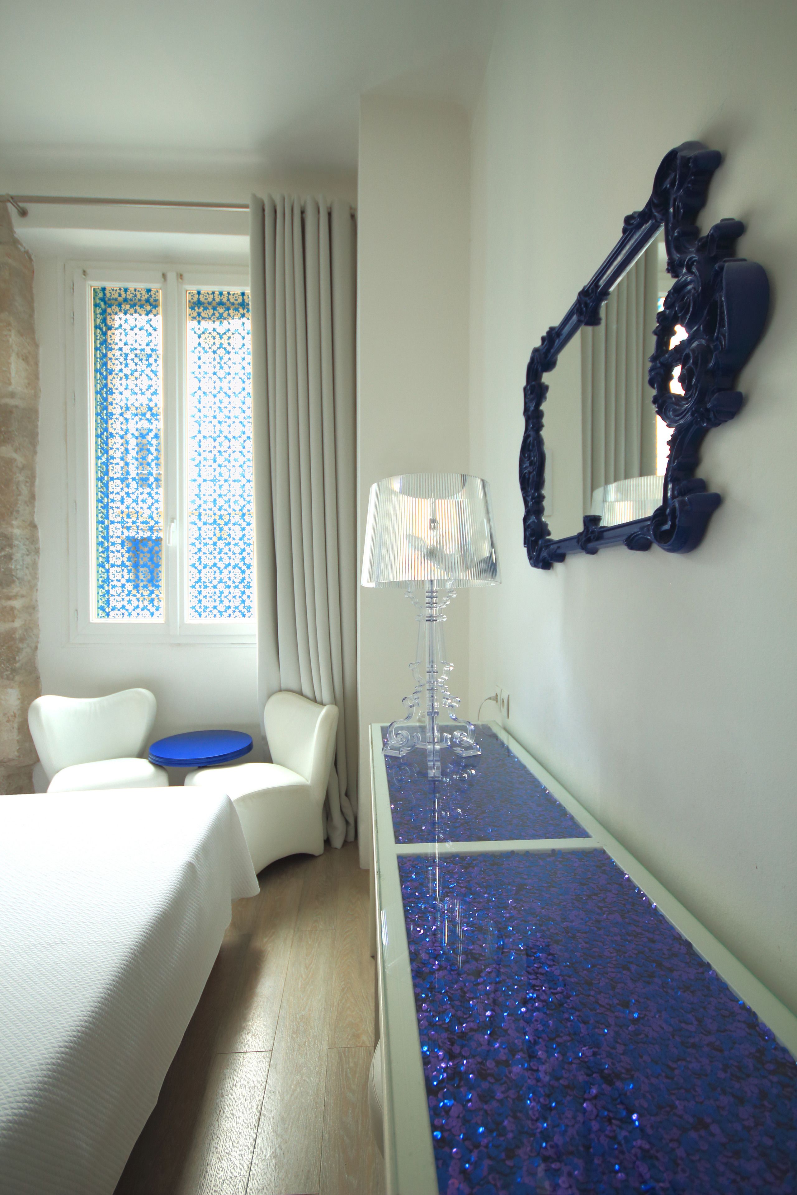 Une chambre d’hôtel avec un lit, une petite table, une chaise, un miroir et une fenêtre avec des rideaux.