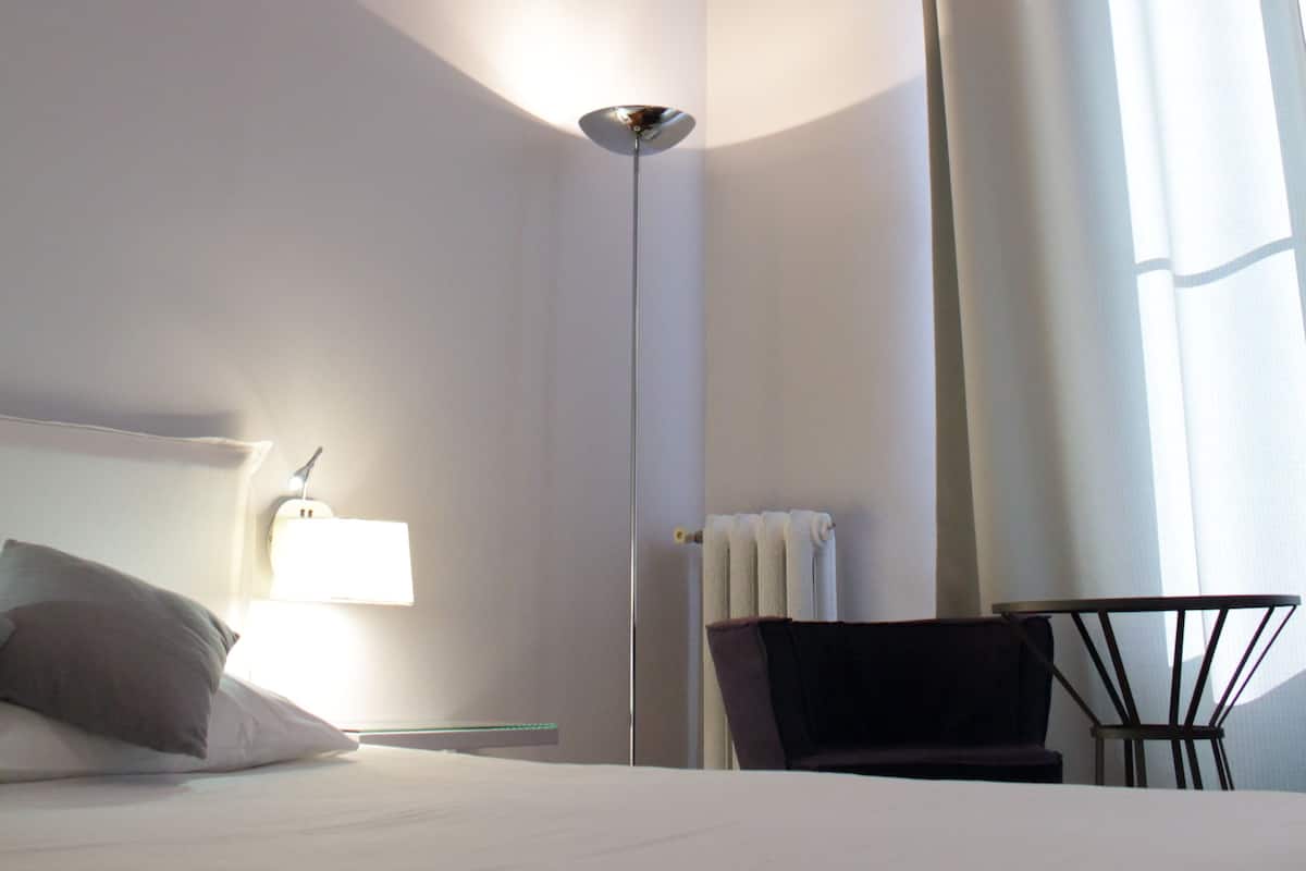 A hotel room with a bed, a nightstand, a lamp, a chair, and a view of the outside through a window with curtains.