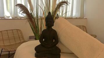 Una stanza con una statua da meditazione su un letto, una poltrona in vimini e un'alta pianta con piume di pampa.