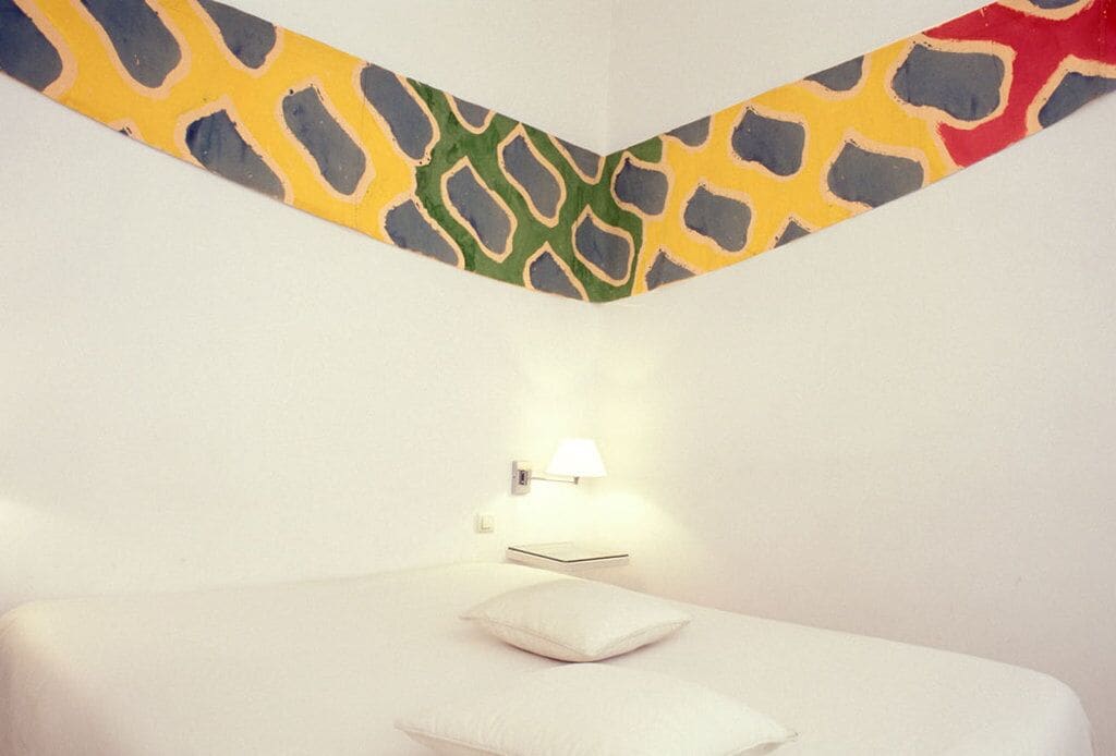 Une chambre à coucher avec un lit, un oreiller, une lampe fixée au mur et un tableau abstrait aux couleurs vives au-dessus du lit.