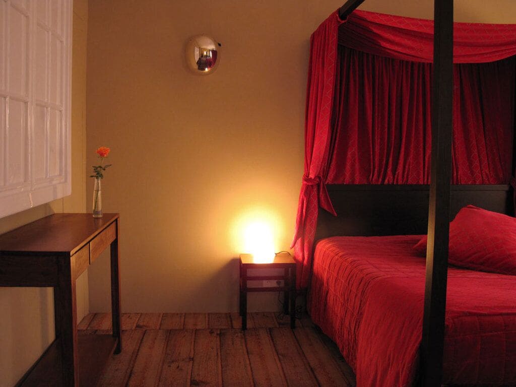 A room with a wooden floor, a bedside table with a vase of flowers, a bed with red bedding, and a wall-mounted light fixture.