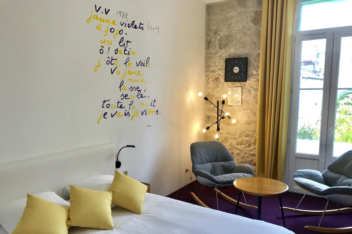 A hotel room with a bed, two yellow pillows, a blue armchair, a small round table, a wall clock, and a wall with handwritten text.
