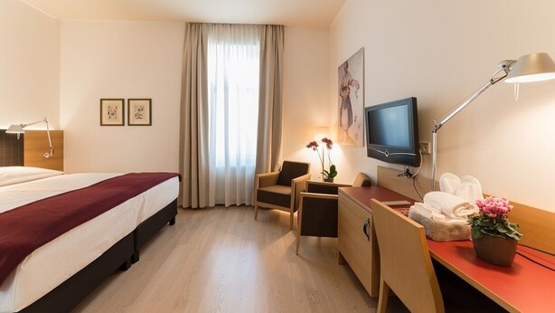 A hotel room with a bed, a desk, a chair, a television, a desk lamp, a potted plant, and a window with curtains.
