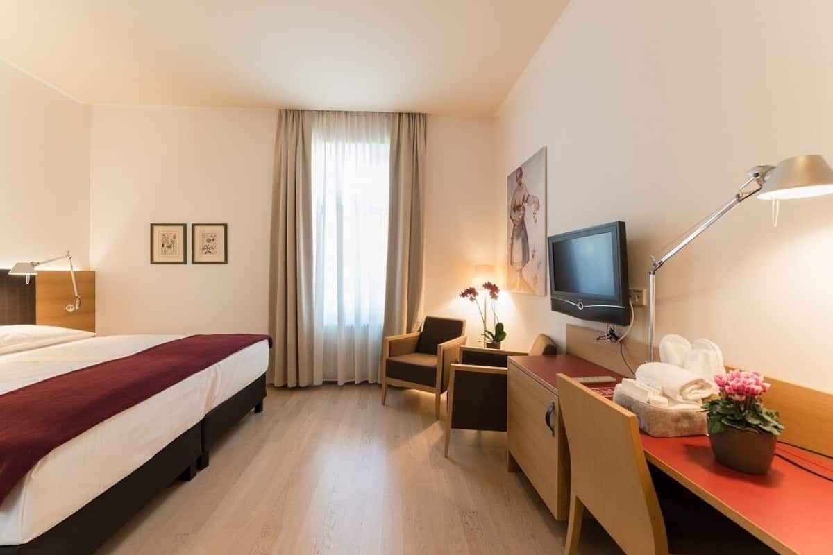 A hotel room with a bed, a desk, a chair, a television, a desk lamp, a potted plant, and a window with curtains.