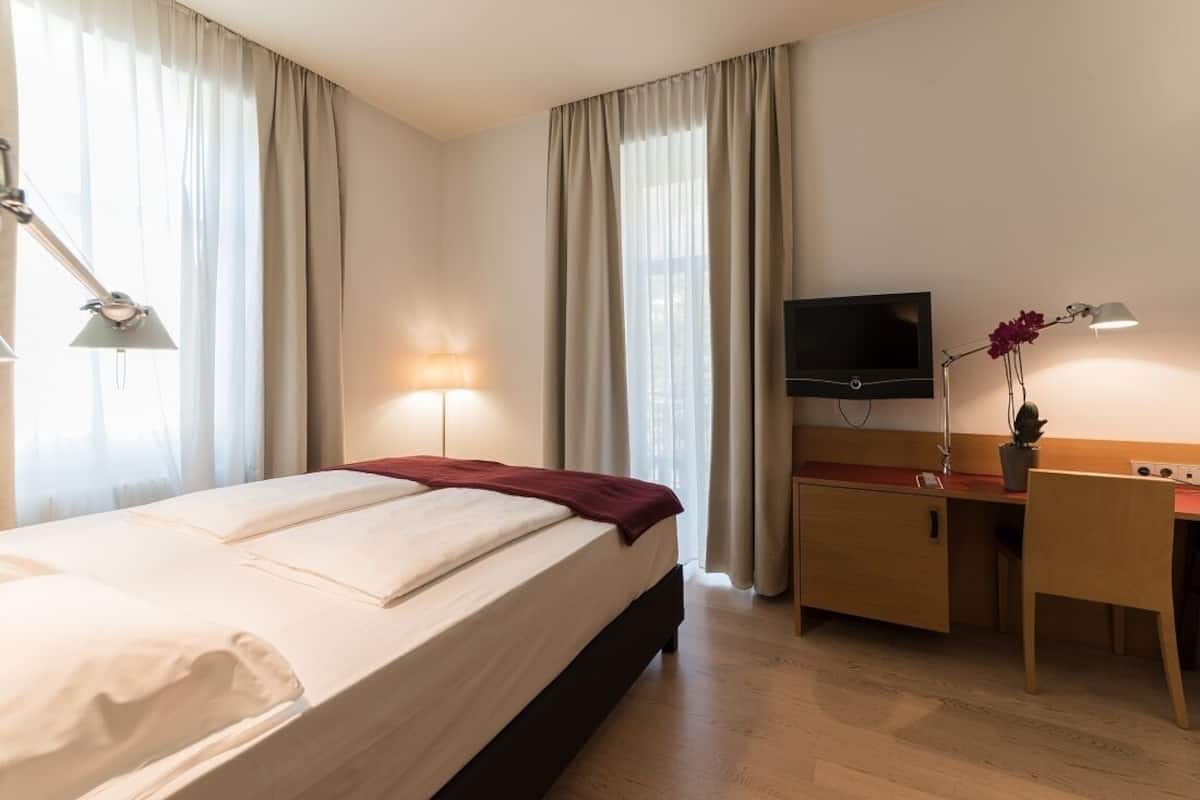A hotel room with a bed, bedside lamp, TV, desk, chair, and a potted plant.