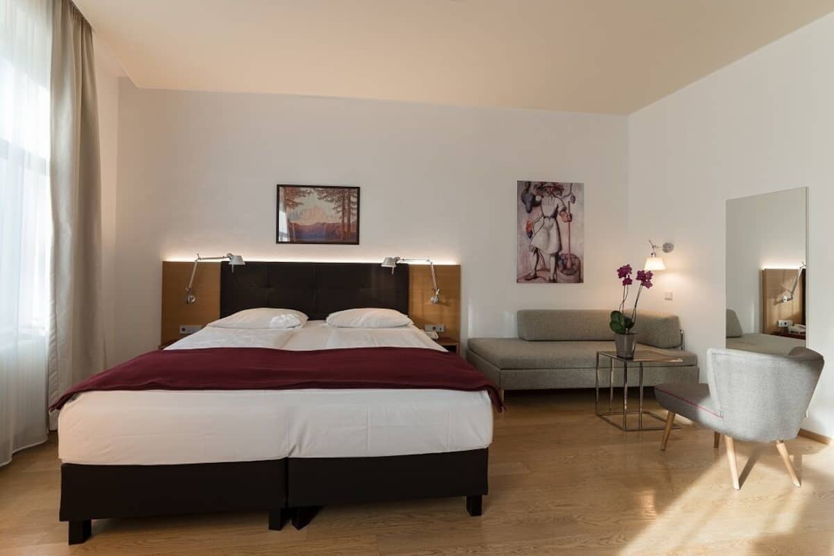 A modern hotel room with a large bed, a grey sofa, a small table with a vase of flowers, and a mirror.