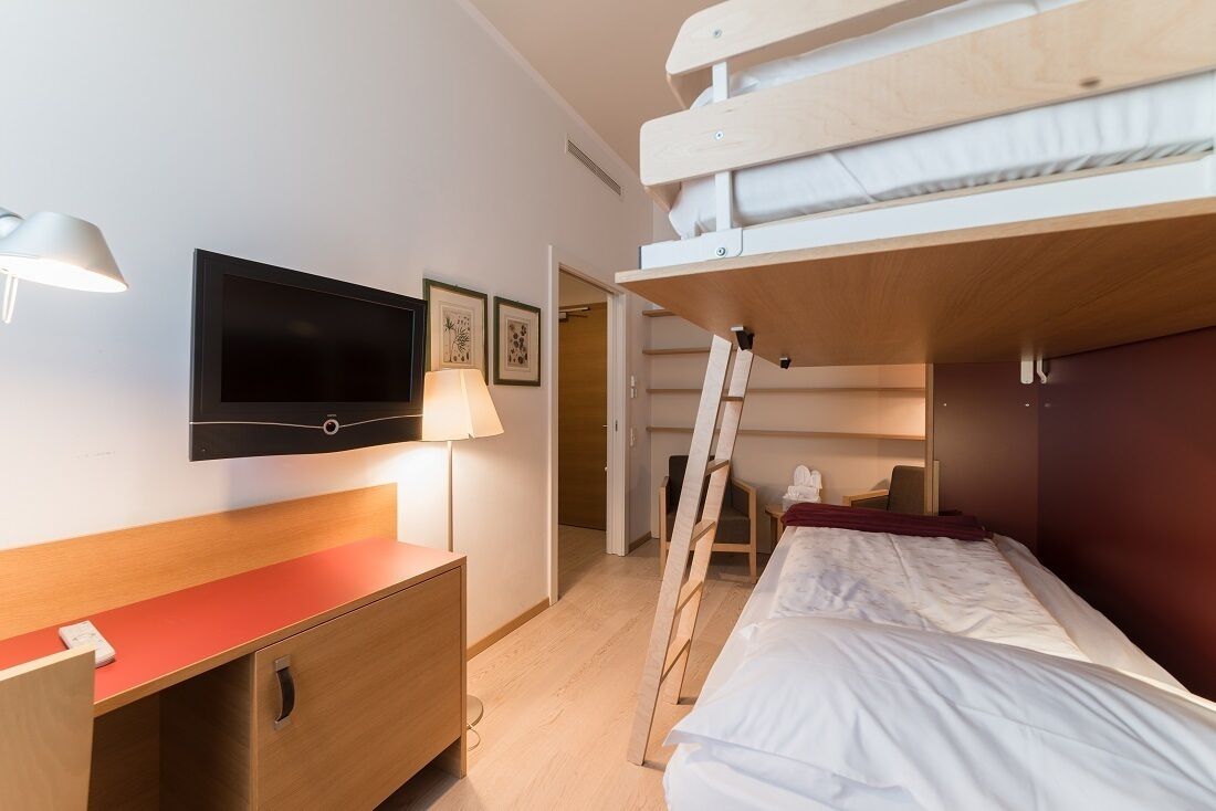 Een compacte hotelkamer met een stapelbed, een bureau met lamp, een flatscreen televisie en ingekaderde foto's aan de muur.