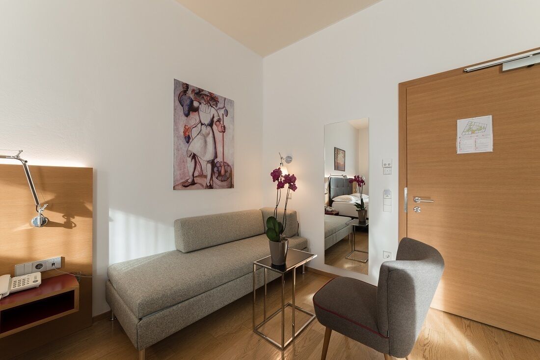 Una habitación de hotel moderna con un sofá gris, una mesita con un florero, un escritorio con una lámpara y una puerta de madera.