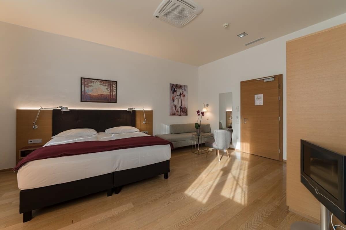 A modern bedroom with a large bed, a bedside table, a small table with a lamp, a sofa, and a TV.