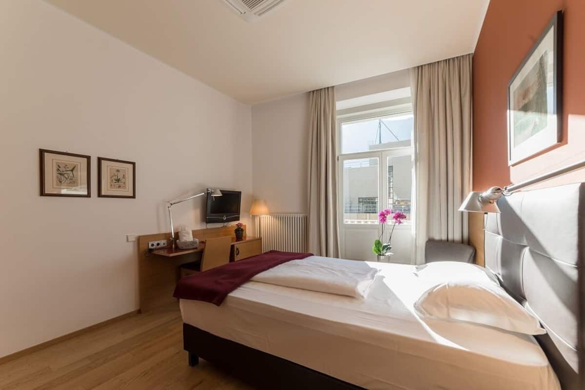 A hotel room with a bed, a desk with a computer, a TV, and a window with curtains.