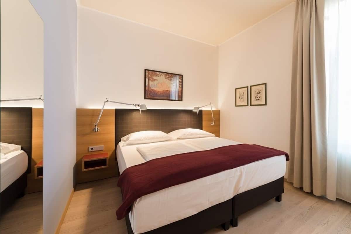 A hotel room with a large bed, bedside tables, a nightstand, a headboard, a painting, and a window with curtains.