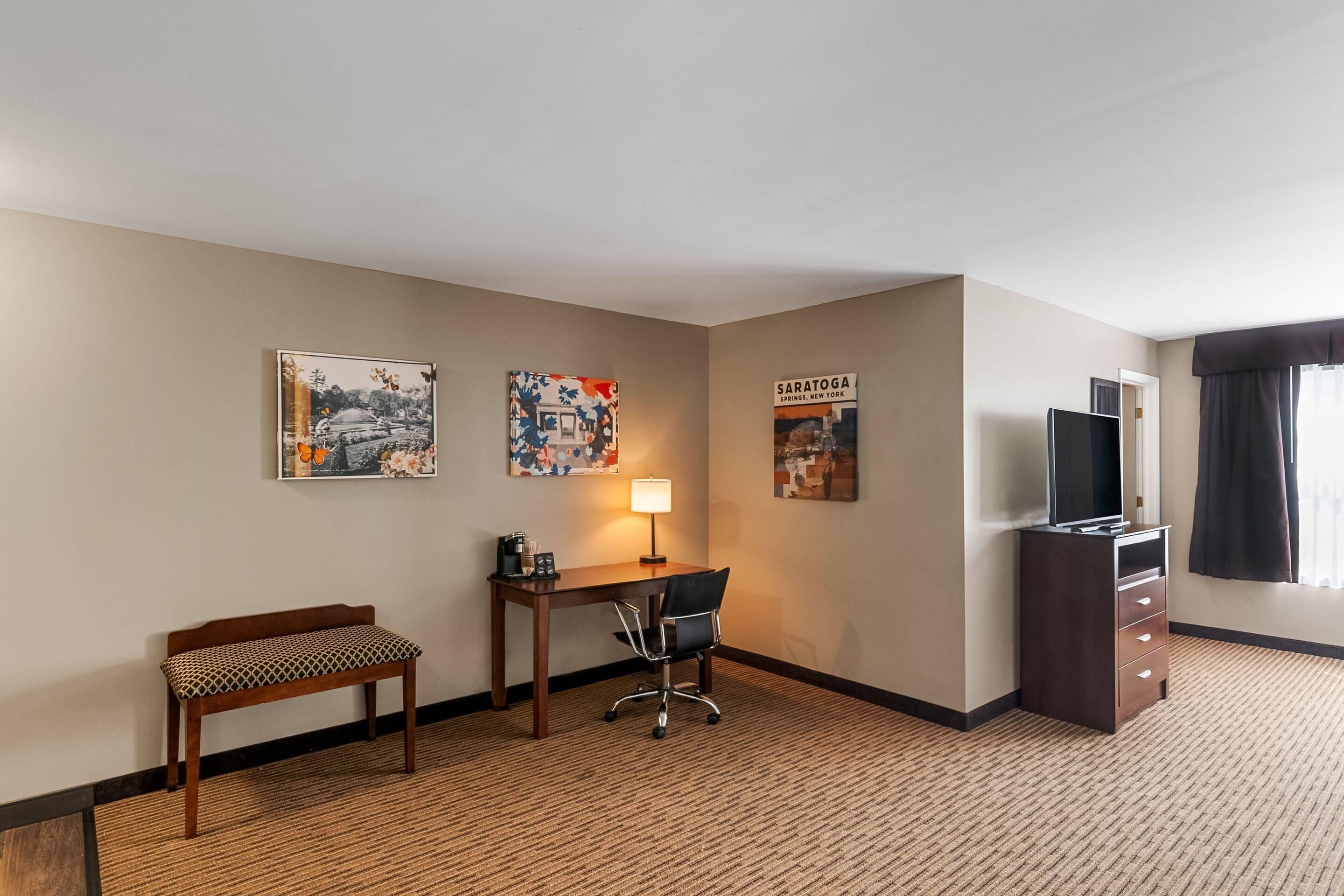 A hotel room with a desk, chair, bench, TV, and artwork on the walls.