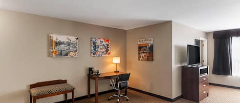 A hotel room with a desk, chair, bench, TV, and artwork on the walls.
