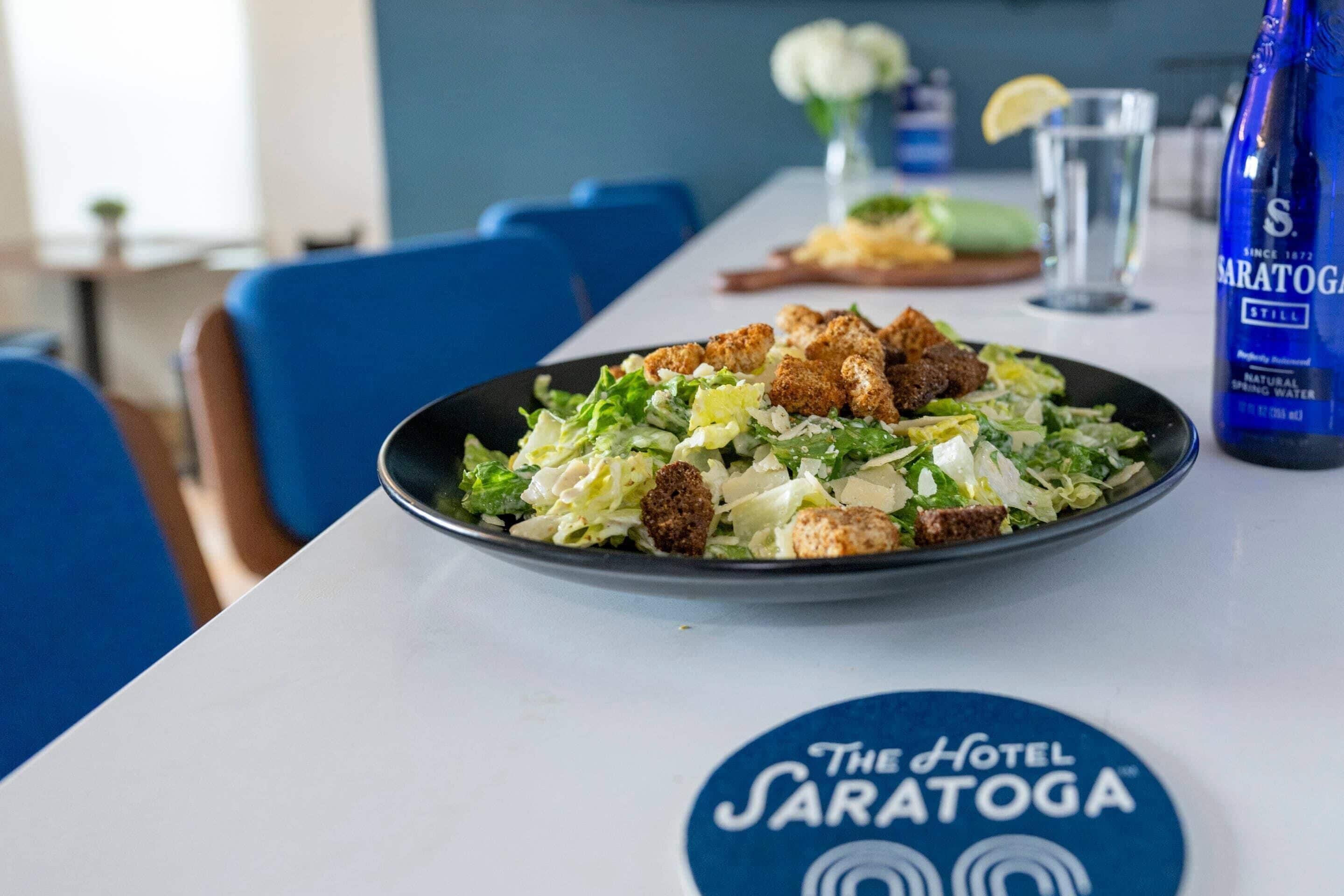 A dining table with a salad, a bottle of water, and a logo for The Hotel Saratoga.