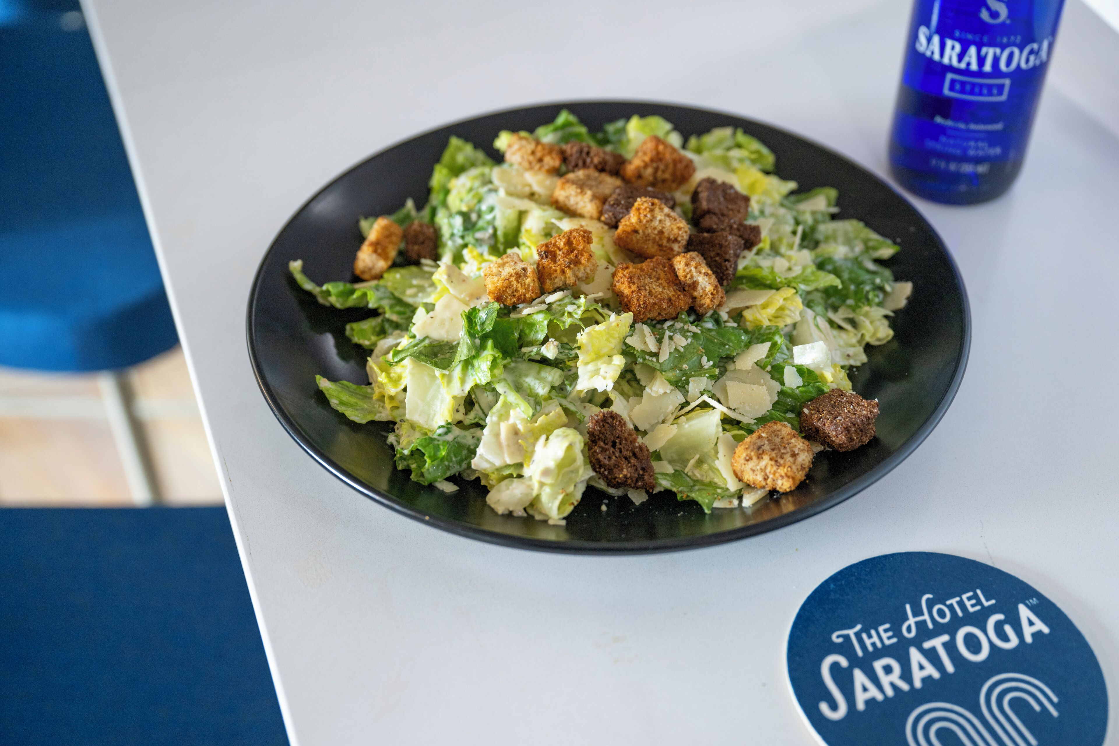 A plate of salad with croutons and cheese on a table with a blue coaster labeled "The Hotel Saratoga."