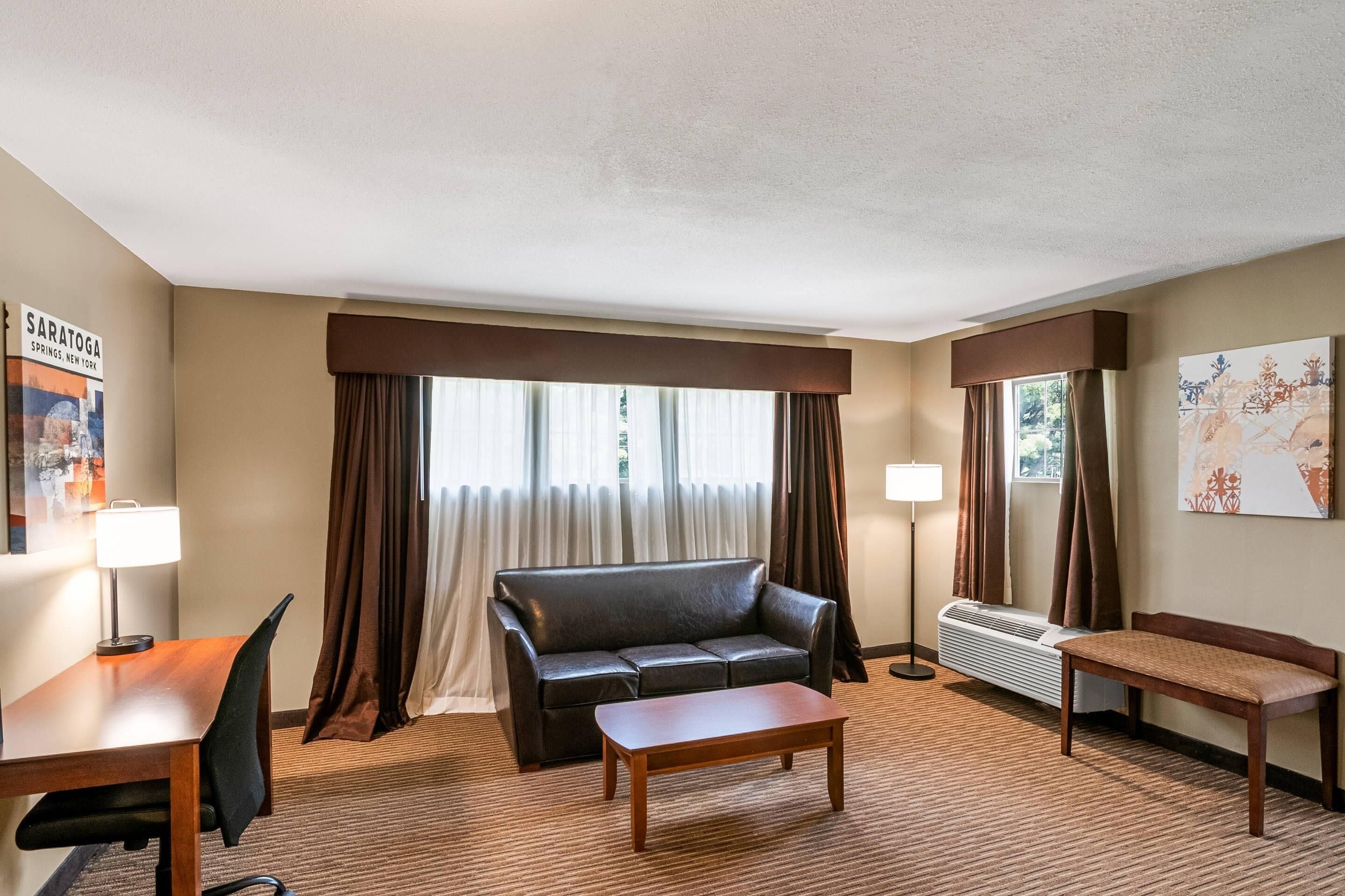 Een hotelkamer met een bank, een klein tafeltje, een bureau en een stoel. Er is een raam met gordijnen, een lamp en een poster aan de muur.