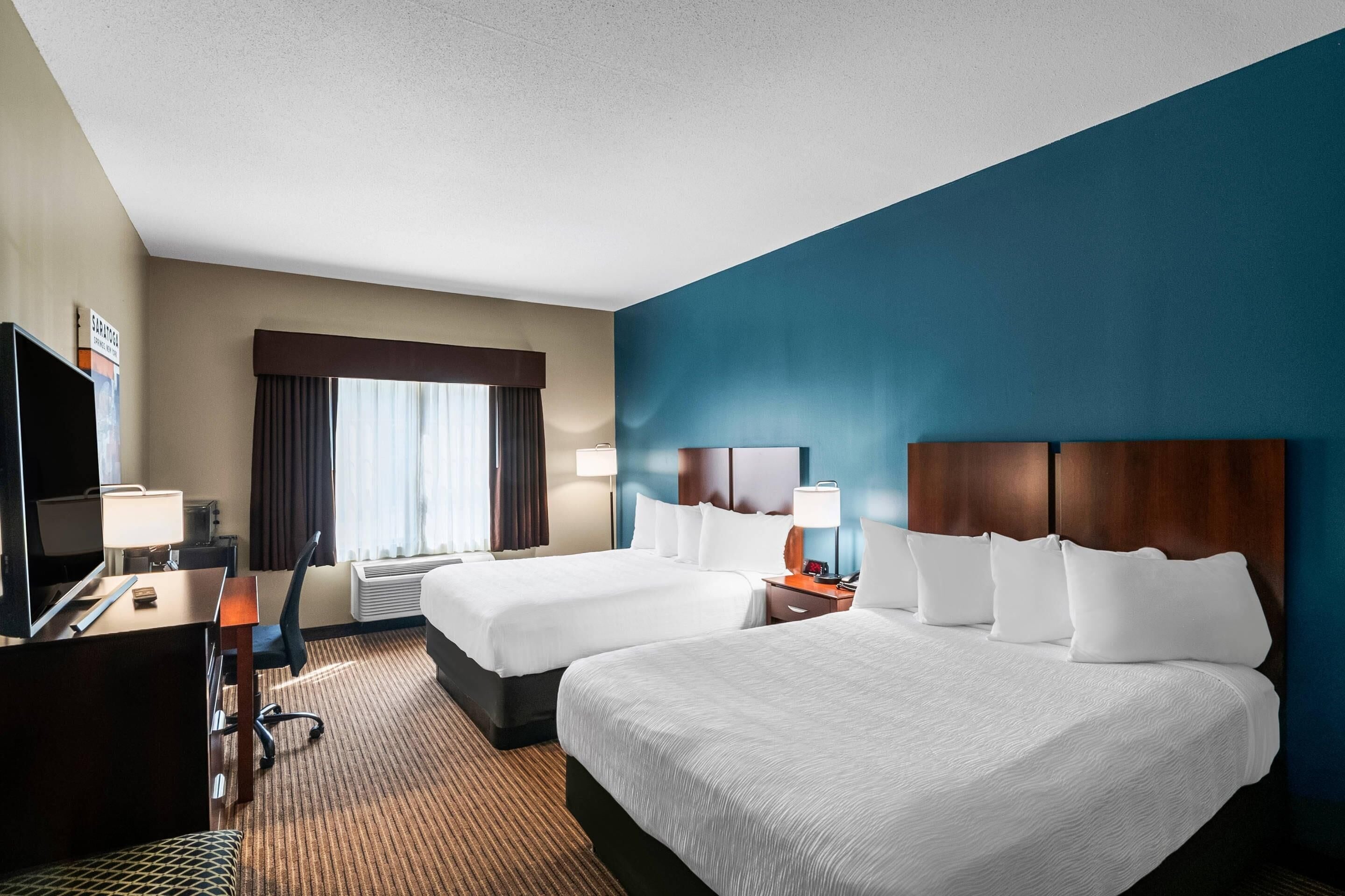 A hotel room with two beds, a desk with a computer, a TV, and a window with curtains.