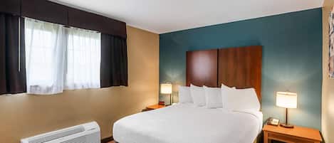 Habitación de hotel con una cama grande, una mesita de noche con lámpara, aire acondicionado instalado en la pared y una ventana con cortinas.