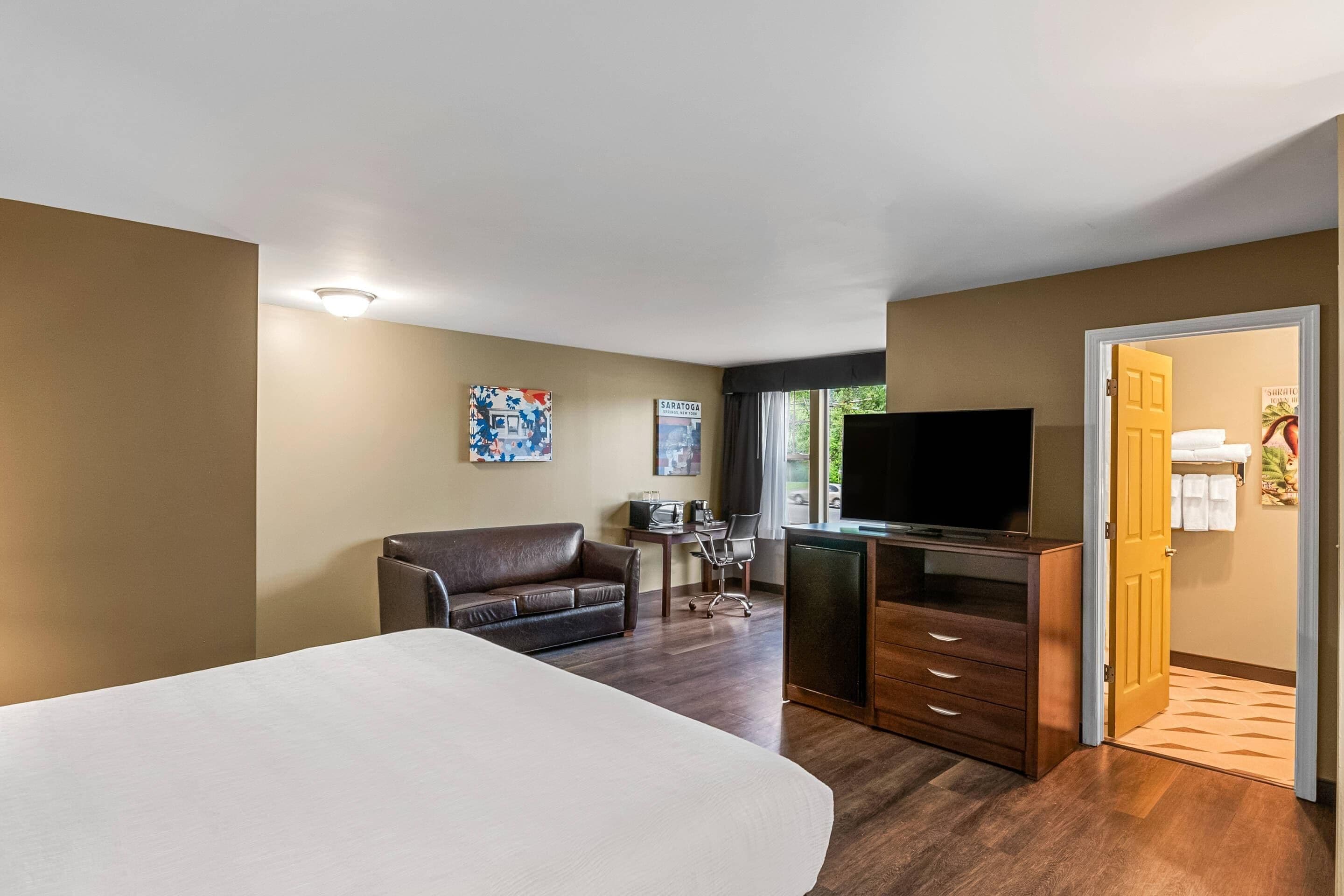 A hotel room with a bed, a desk, a television, a sofa, and a door leading to a bathroom.