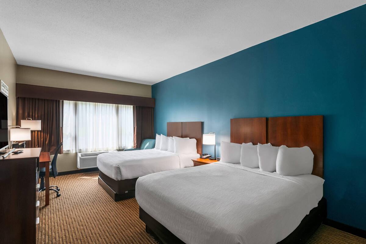 A hotel room with two beds, a desk, a chair, a desk lamp, and a window with curtains.