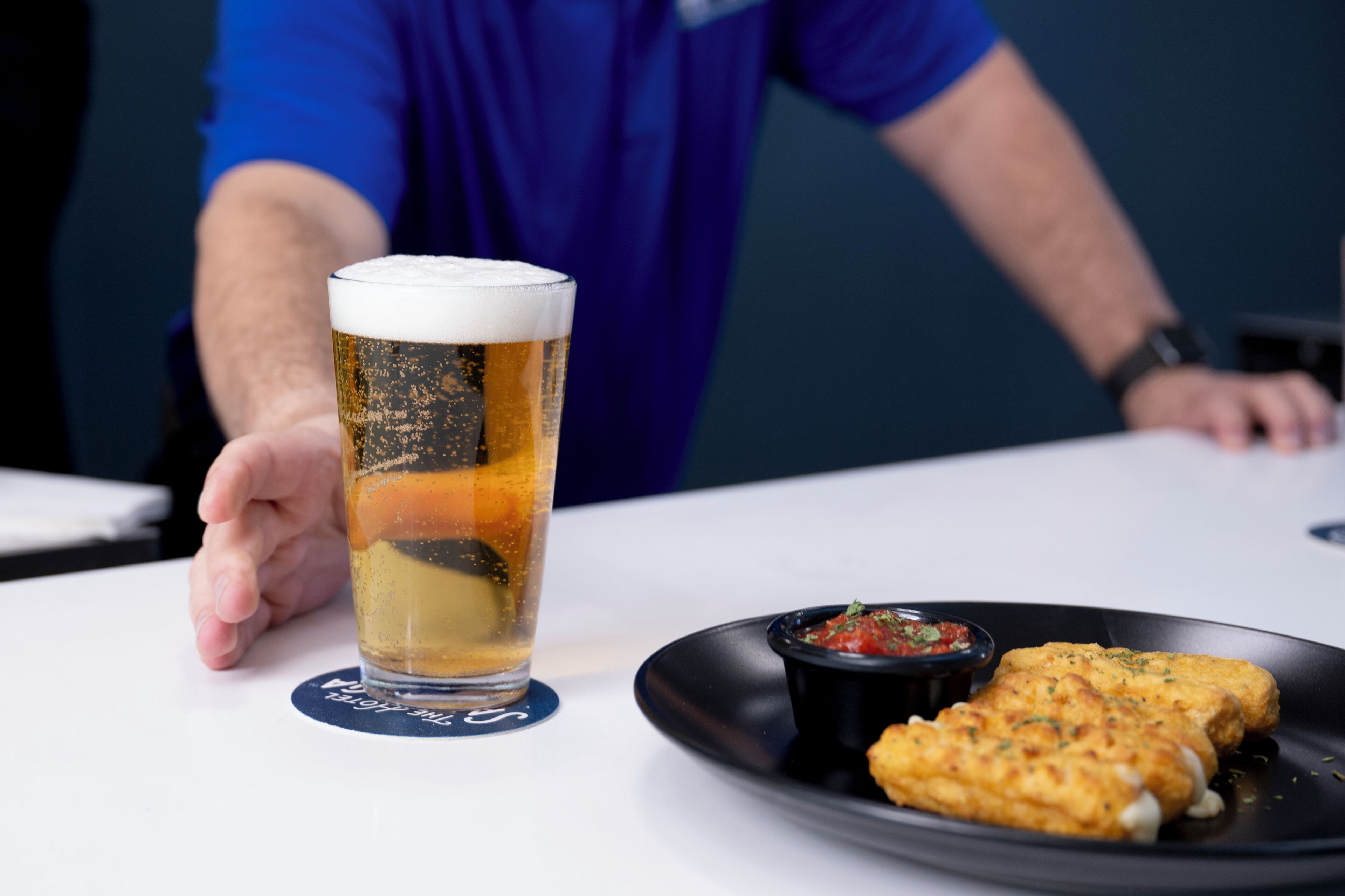 A glass of beer on a coaster, a plate with two pieces of food, and a person in a blue shirt.