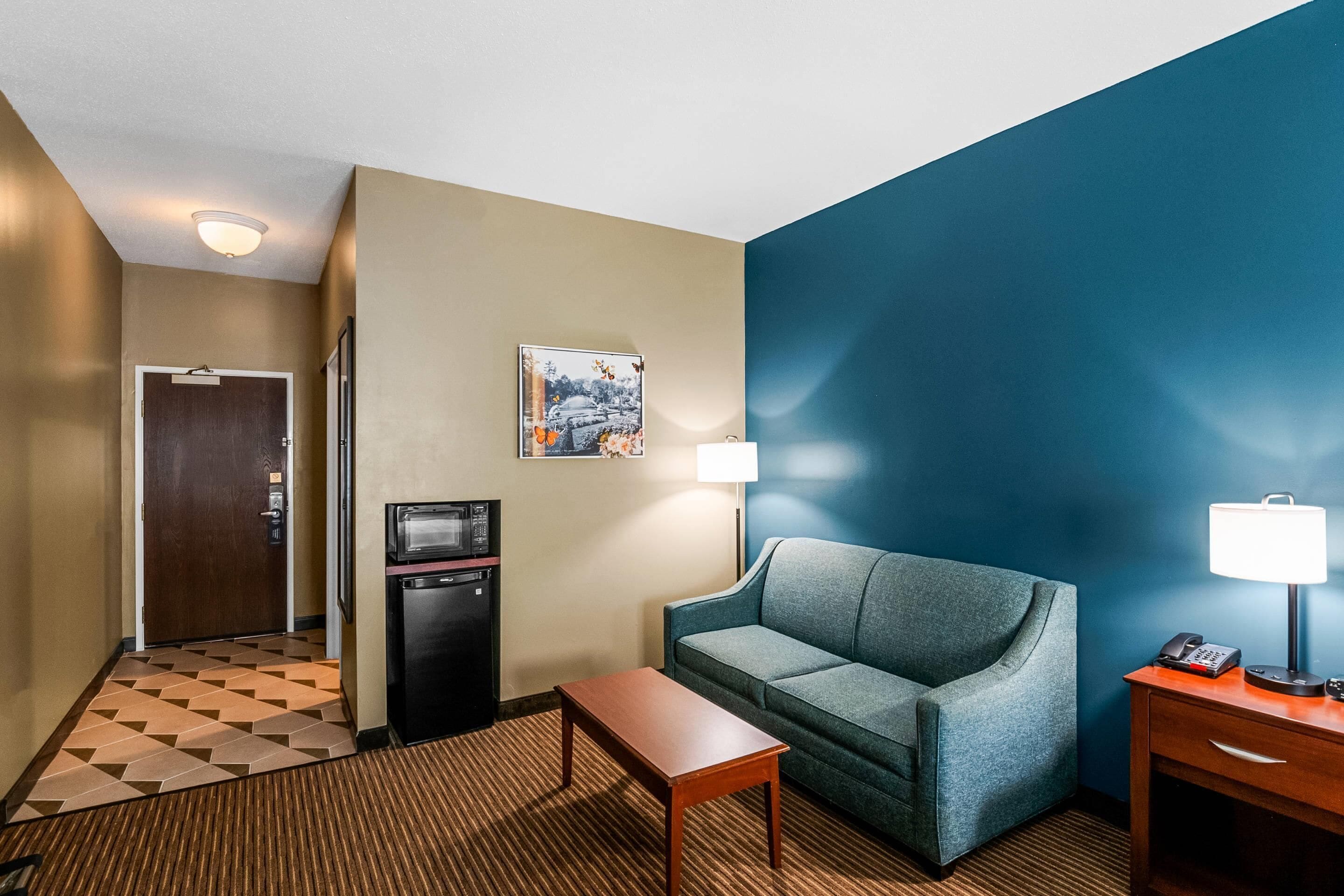 A hotel room with a check-in counter, a microwave, a small refrigerator, a sofa, a table, a lamp, and a painting on the wall.