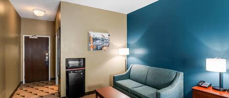 Habitación de hotel con mostrador de recepción, microondas, refrigerador pequeño, sofá, mesa, lámpara y un cuadro en la pared.