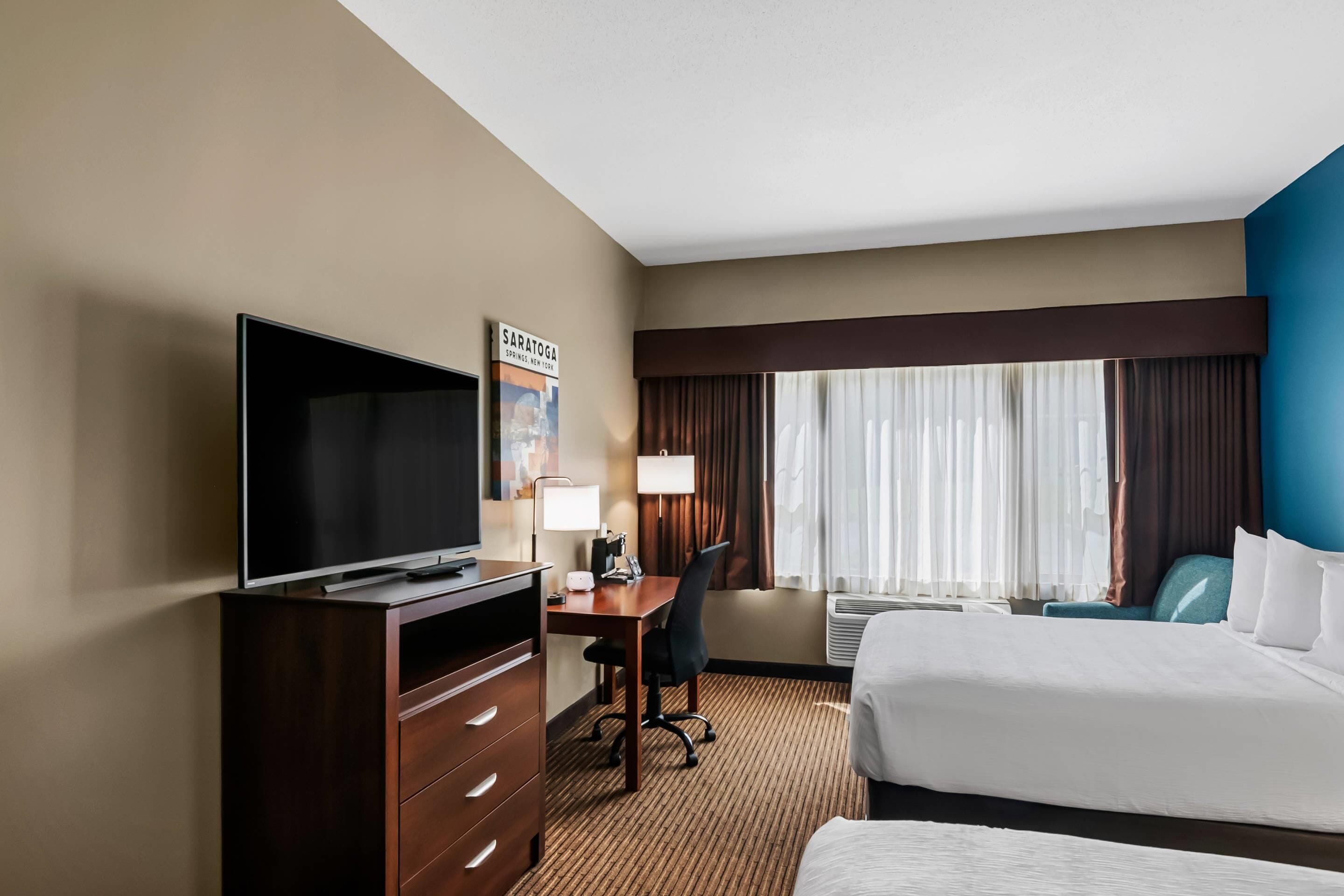 A hotel room with a flat-screen TV, a desk with a chair, a bed, and a window with curtains.