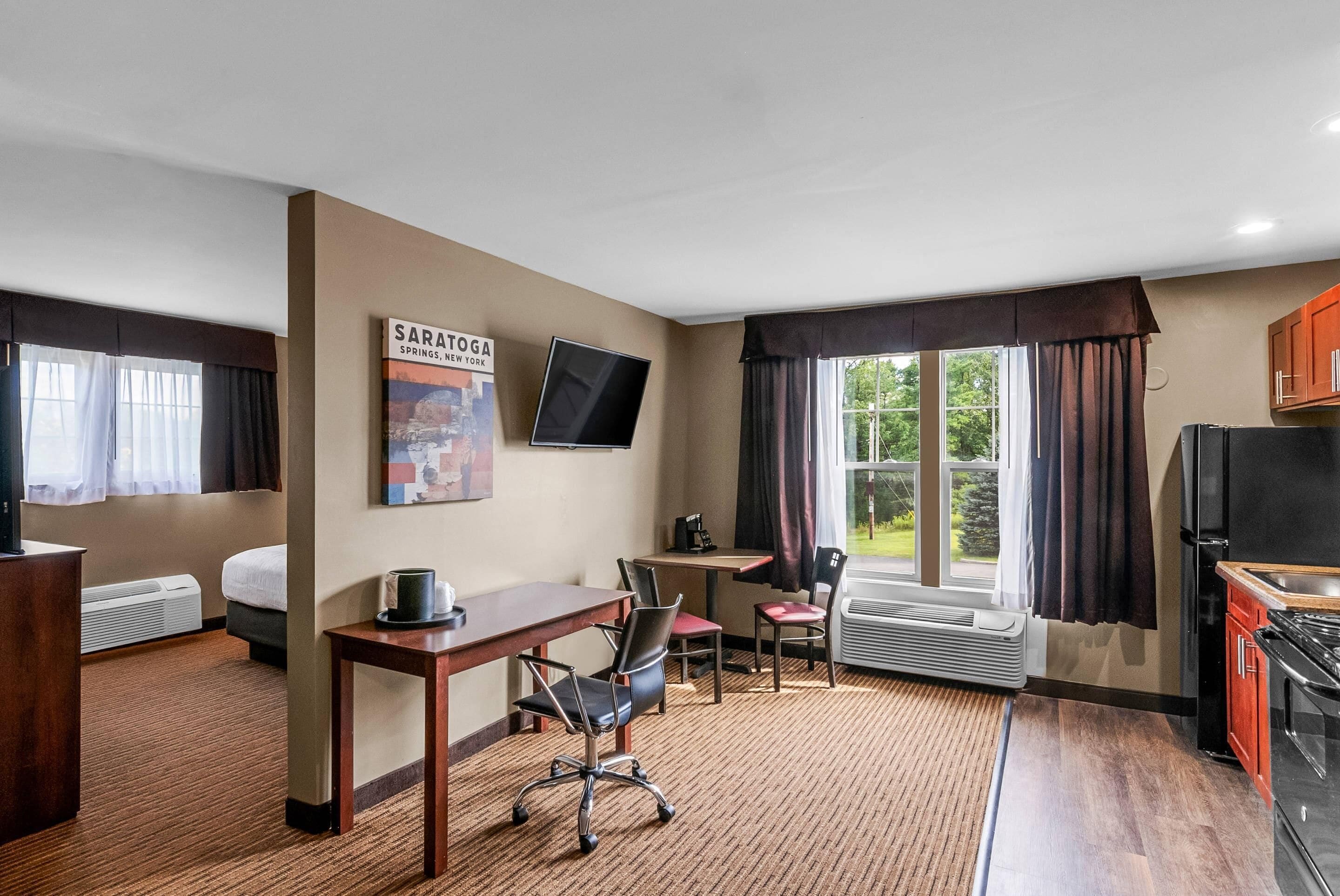 A hotel room with a bed, a desk, a chair, a TV, a window with curtains, and a kitchen area.
