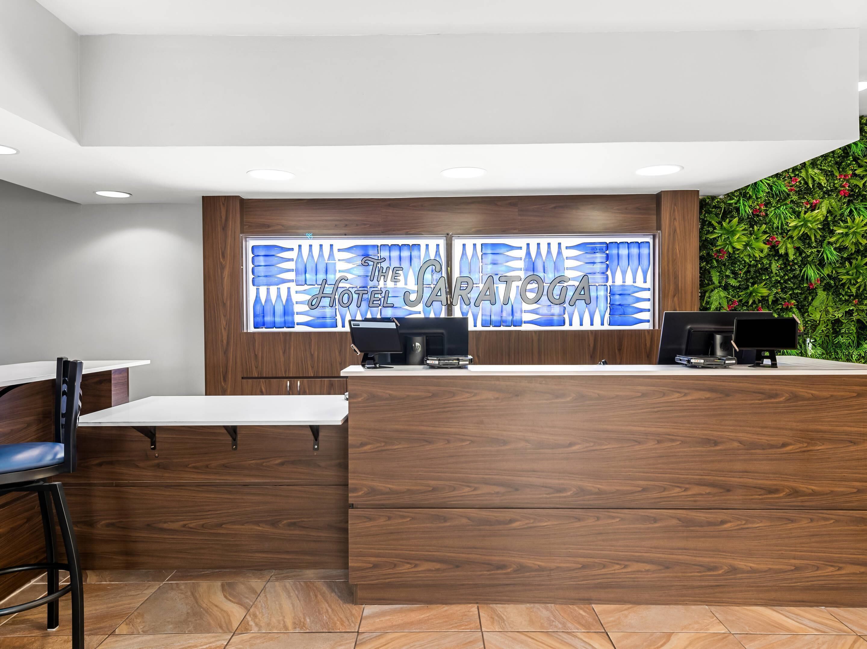 Una recepción con detalles de madera, un muro de cristal con motivos azules tipo botella y una pared verde cubierta de plantas.