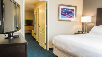 Une chambre d’hôtel avec un lit, un téléviseur posé sur une commode, un bureau, une chaise, une porte donnant sur une autre pièce et un tableau encadré accroché au mur.