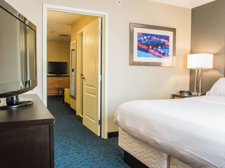Une chambre d’hôtel avec un lit, un téléviseur posé sur une commode, un bureau, une chaise, une porte donnant sur une autre pièce et un tableau encadré accroché au mur.