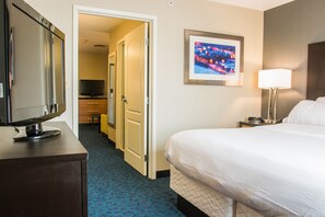 A hotel room with a bed, a TV on a dresser, a desk with a lamp, and a door leading to another room.