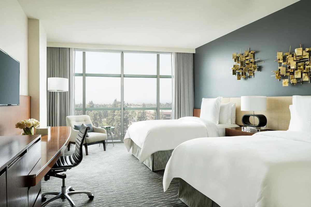 Ein Hotelzimmer mit zwei Betten, einem Schreibtisch, einem Sessel, einem Fernseher und Blick auf die Stadt.
