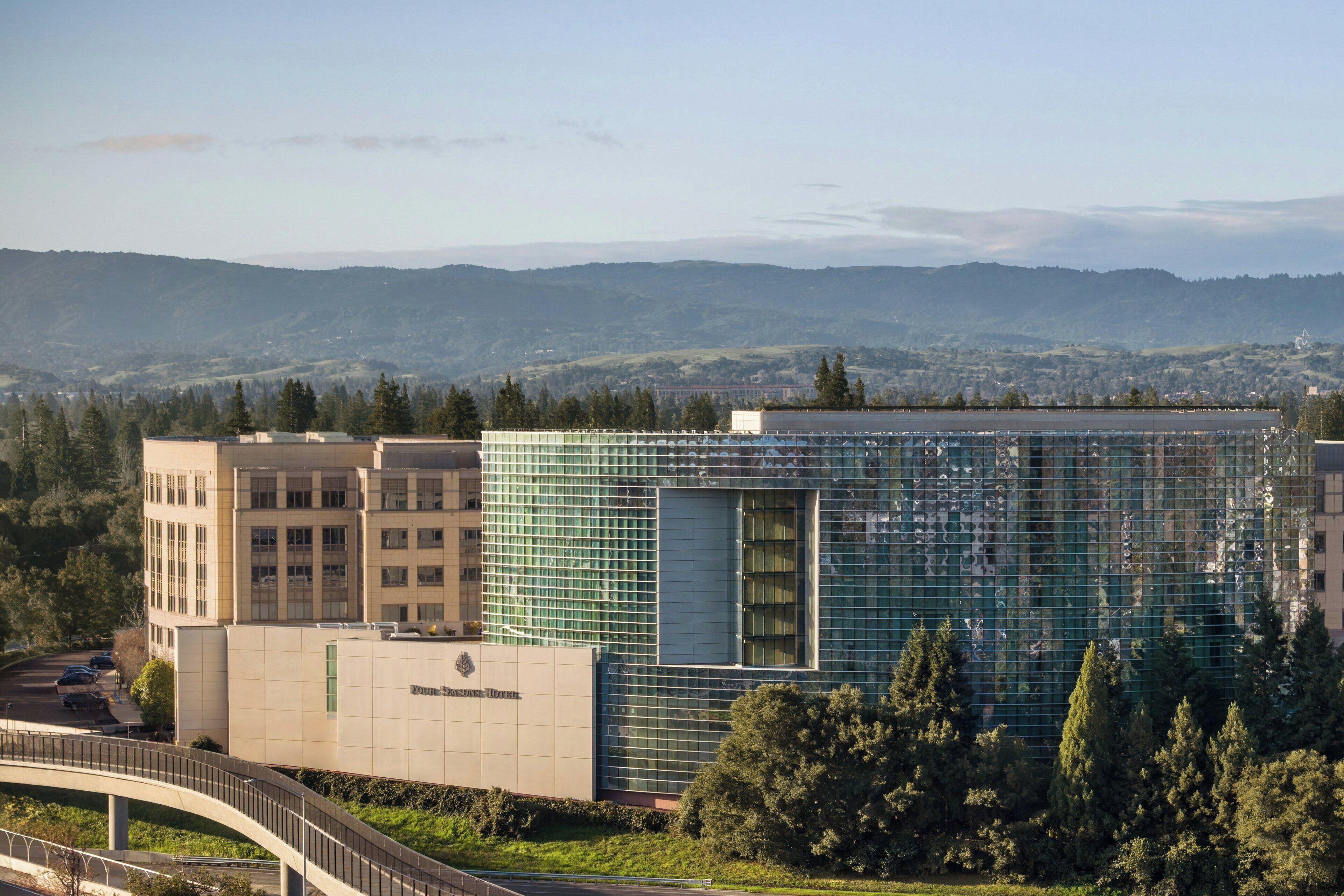 Un bâtiment moderne à la façade de verre, entouré de verdure et traversé par un pont.