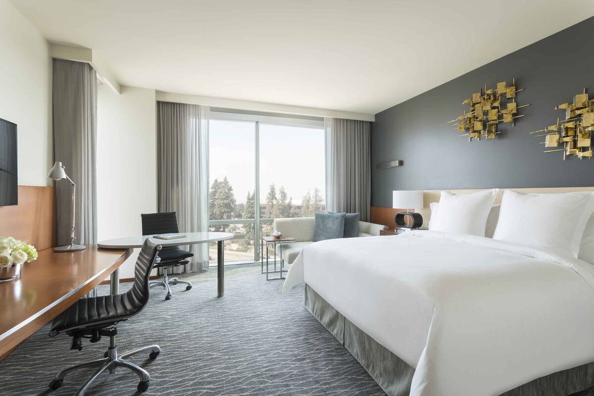 Ein Hotelzimmer mit einem großen Bett, einem Schreibtisch mit Stuhl, einem kleinen Tisch und einer Couch. Es gibt ein großes Fenster mit Vorhängen, einen Flachbildfernseher und eine Wanddekoration.