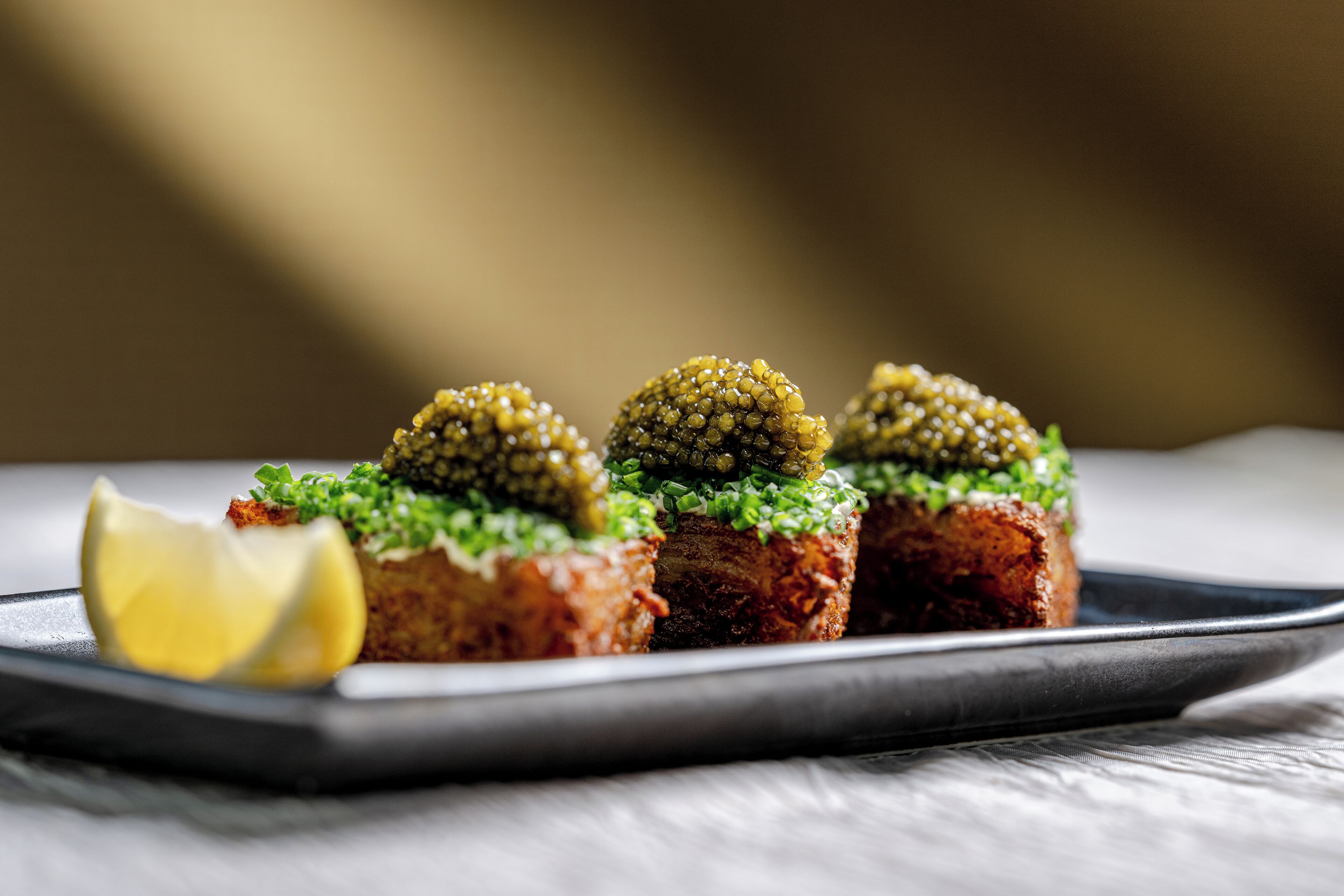 Une assiette contenant trois portions de nourriture, surmontées de caviar vert et garnies d’oignons verts, accompagnée d’un quartier de citron.