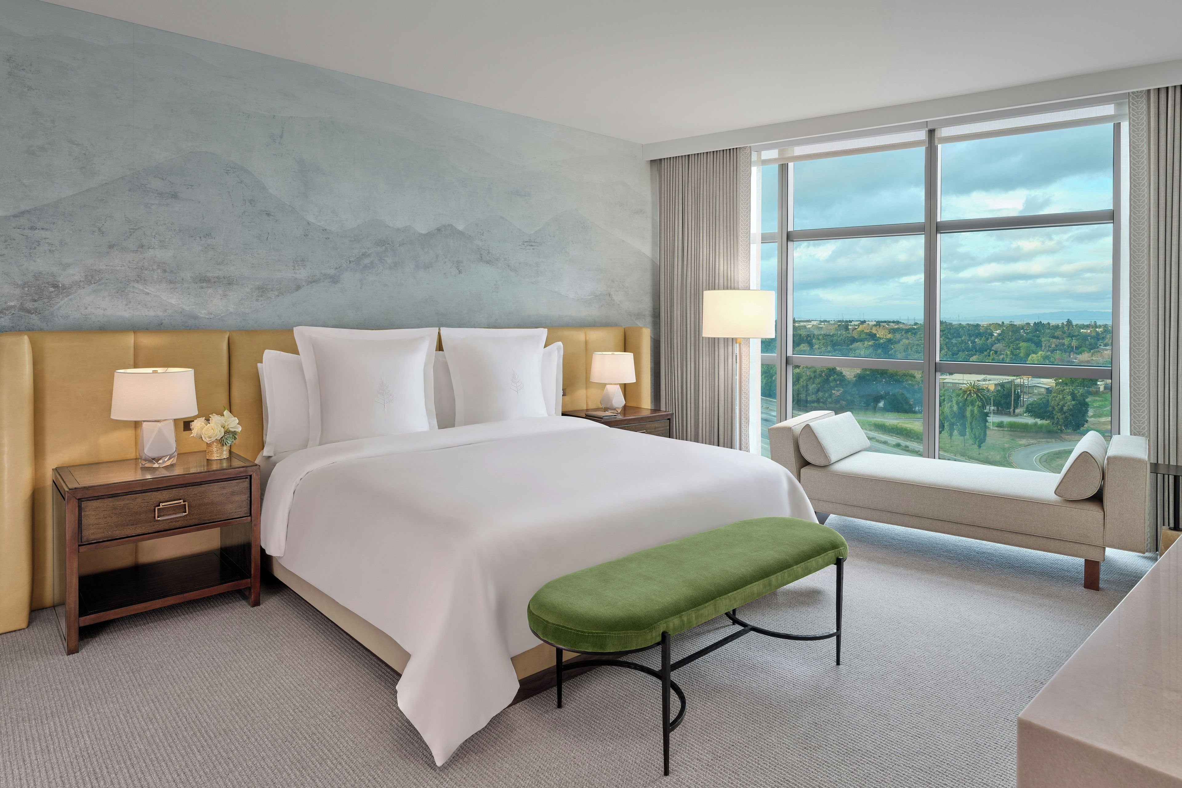 Quarto de hotel com uma cama grande, mesa de cabeceira com luminária, um banco verde e vista para a cidade através de grandes janelas.