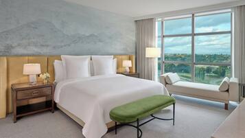 Quarto de hotel com uma cama grande, mesa de cabeceira com luminária, um banco verde e vista para a cidade através de grandes janelas.