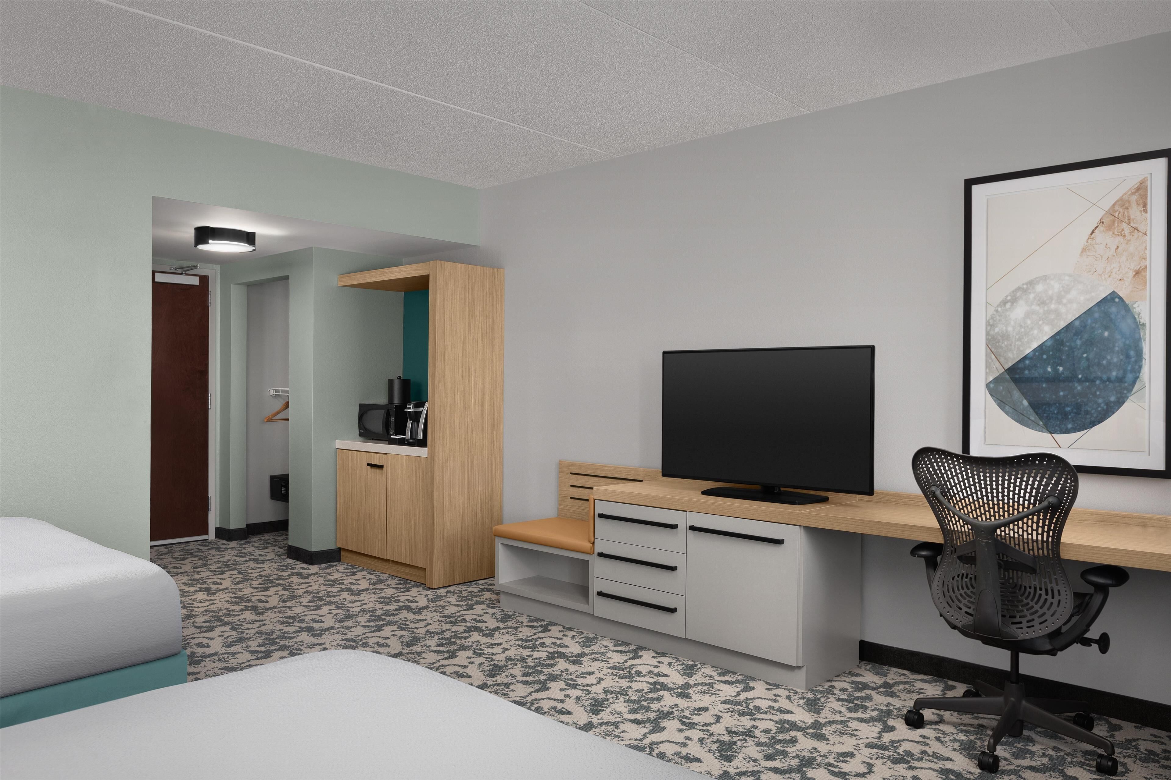 A hotel room with a bed, a desk with a computer, a chair, a TV, and a framed artwork on the wall.
