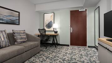Quarto de hotel com sofá, escrivaninha com luminária, televisão e porta.