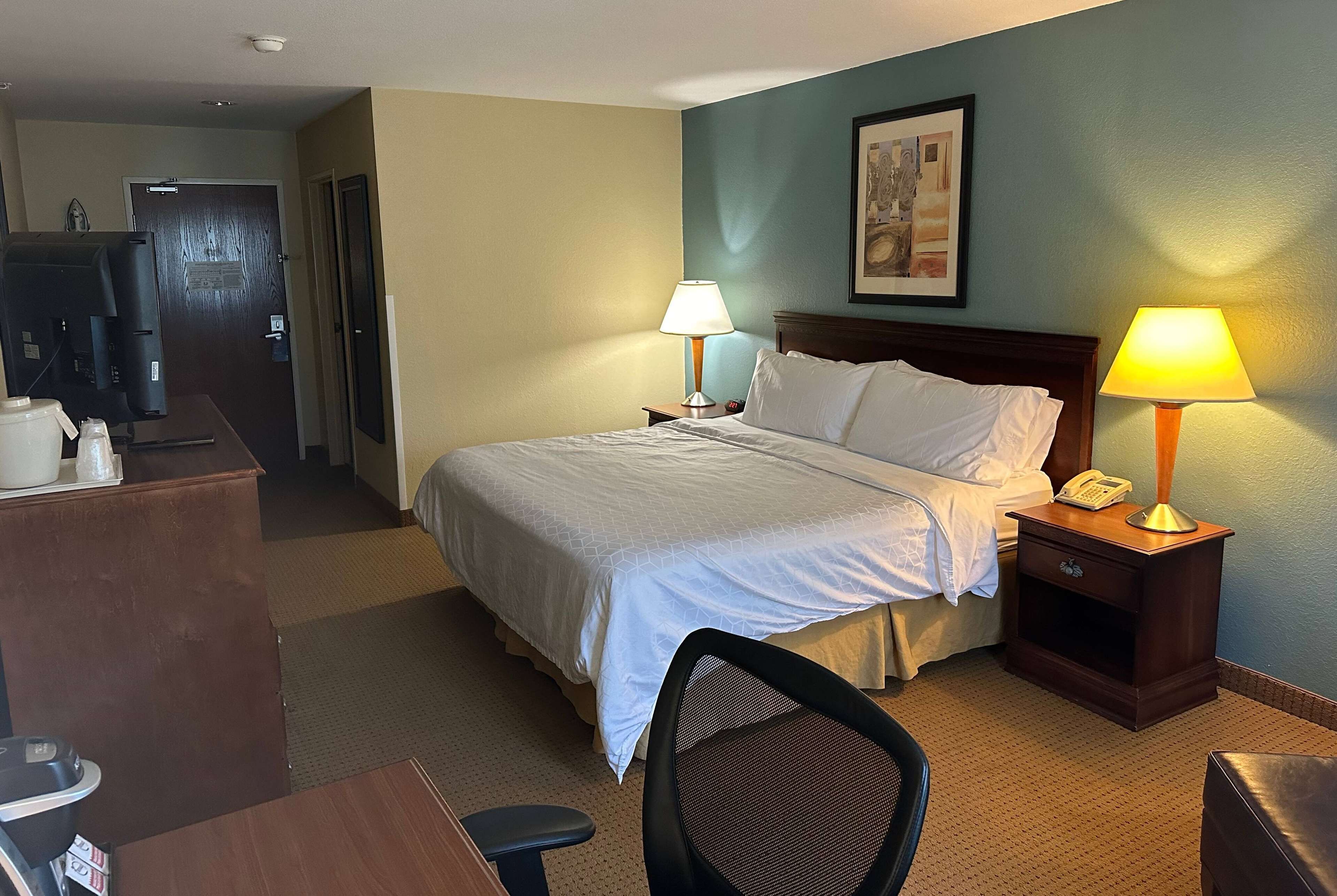 A hotel room with a bed, two bedside lamps, a desk with a computer, a chair, and a small table with a pitcher and glasses.