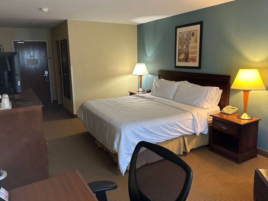 A hotel room with a bed, two bedside lamps, a desk with a computer, a chair, and a small table with a pitcher and glasses.
