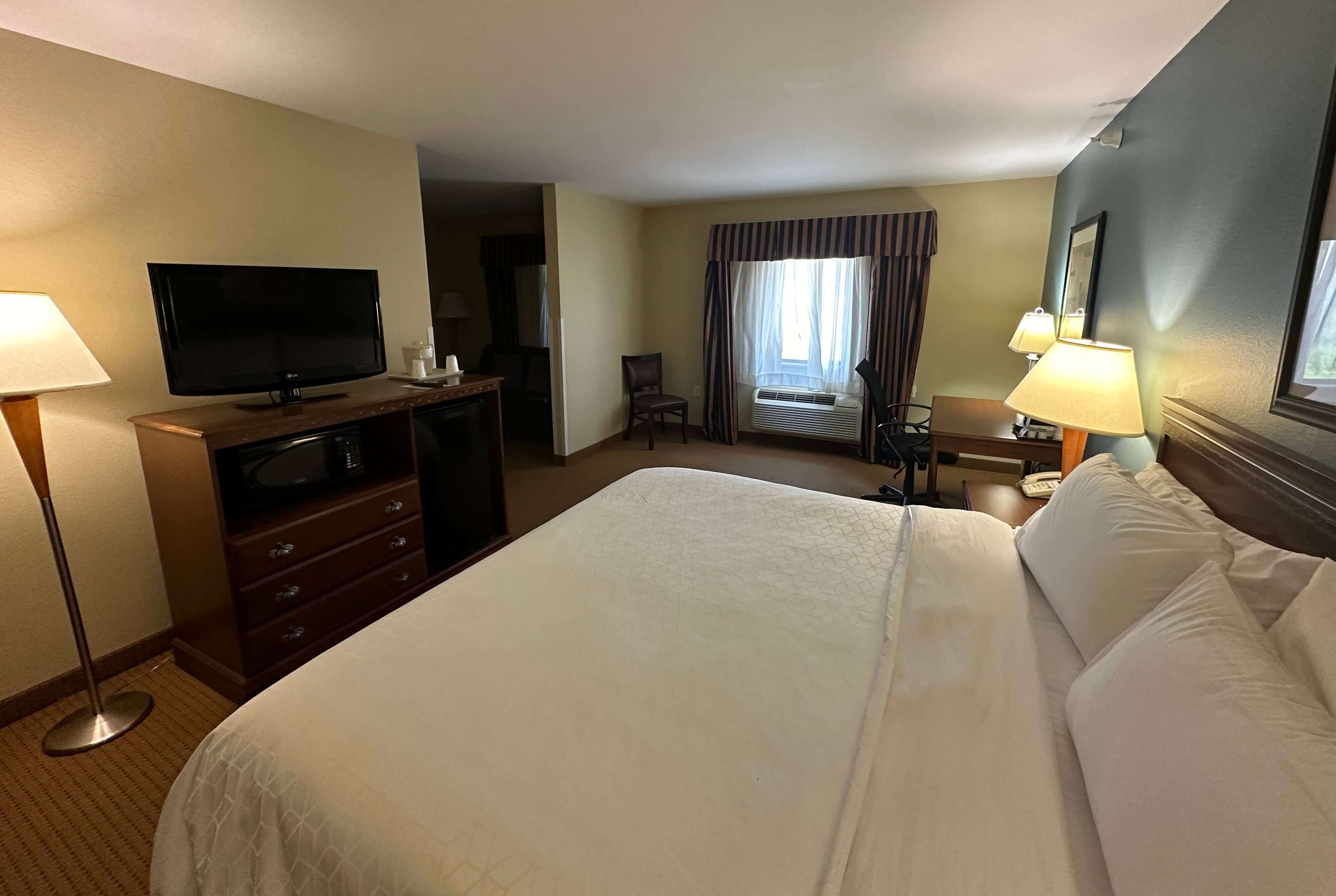 Habitación de hotel con una cama grande, un televisor de pantalla plana, un escritorio con lámpara y una ventana con cortinas.