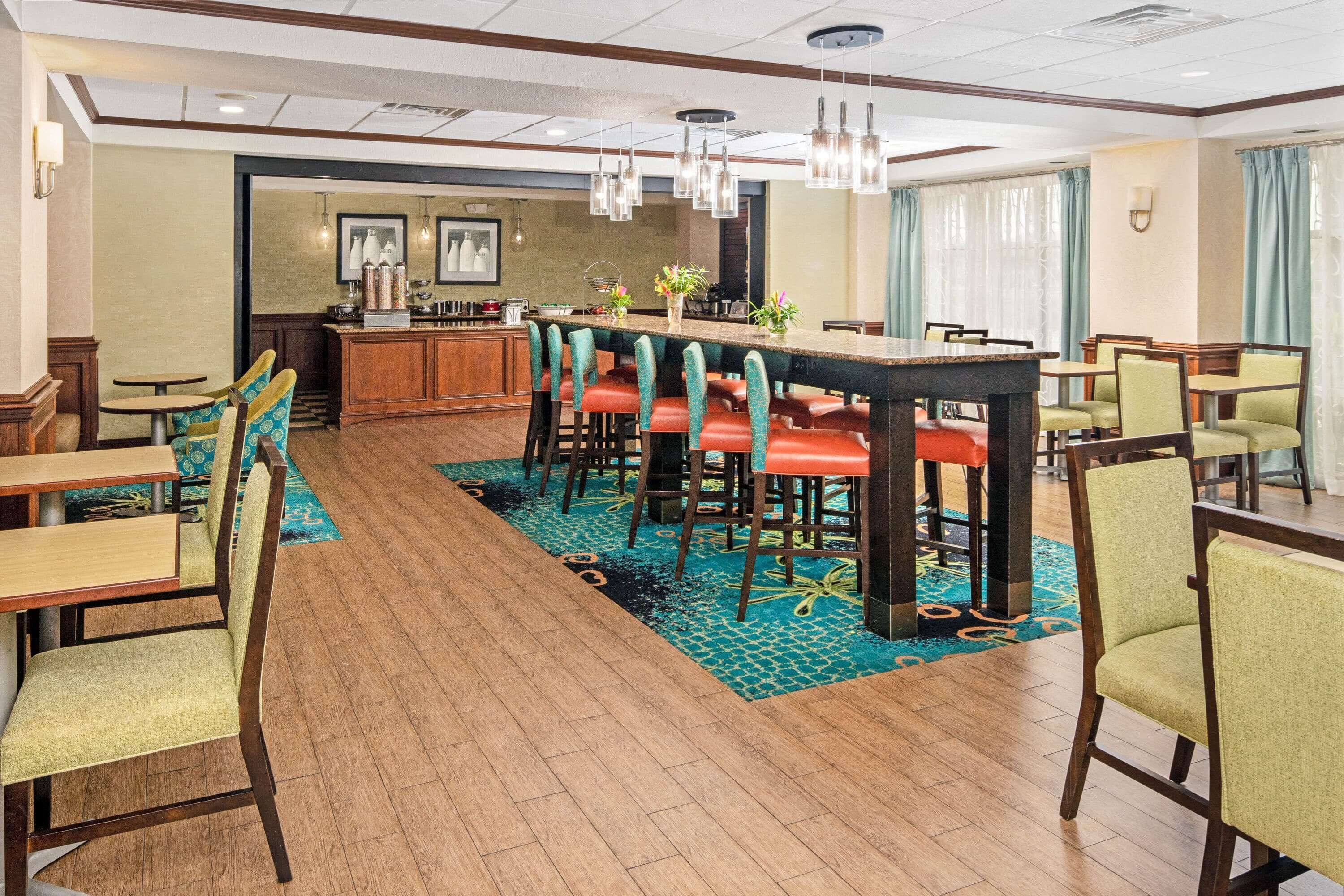 A hotel dining area with a long communal table, multiple bar stools, and several individual tables.