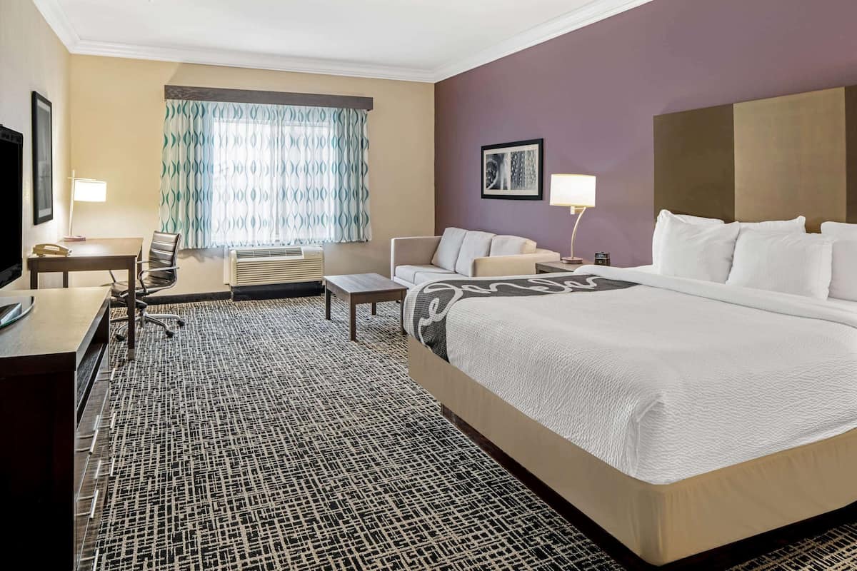 A hotel room with a large bed, a desk with a chair, a sofa, a TV, and a window with curtains.