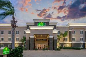 A La Quinta Inn & Suites building with a large entrance, palm trees, and a sunset sky. - La Quinta Inn & Suites by Wyndham Corpus Christi-N Padre Isl (Corpus Christi)