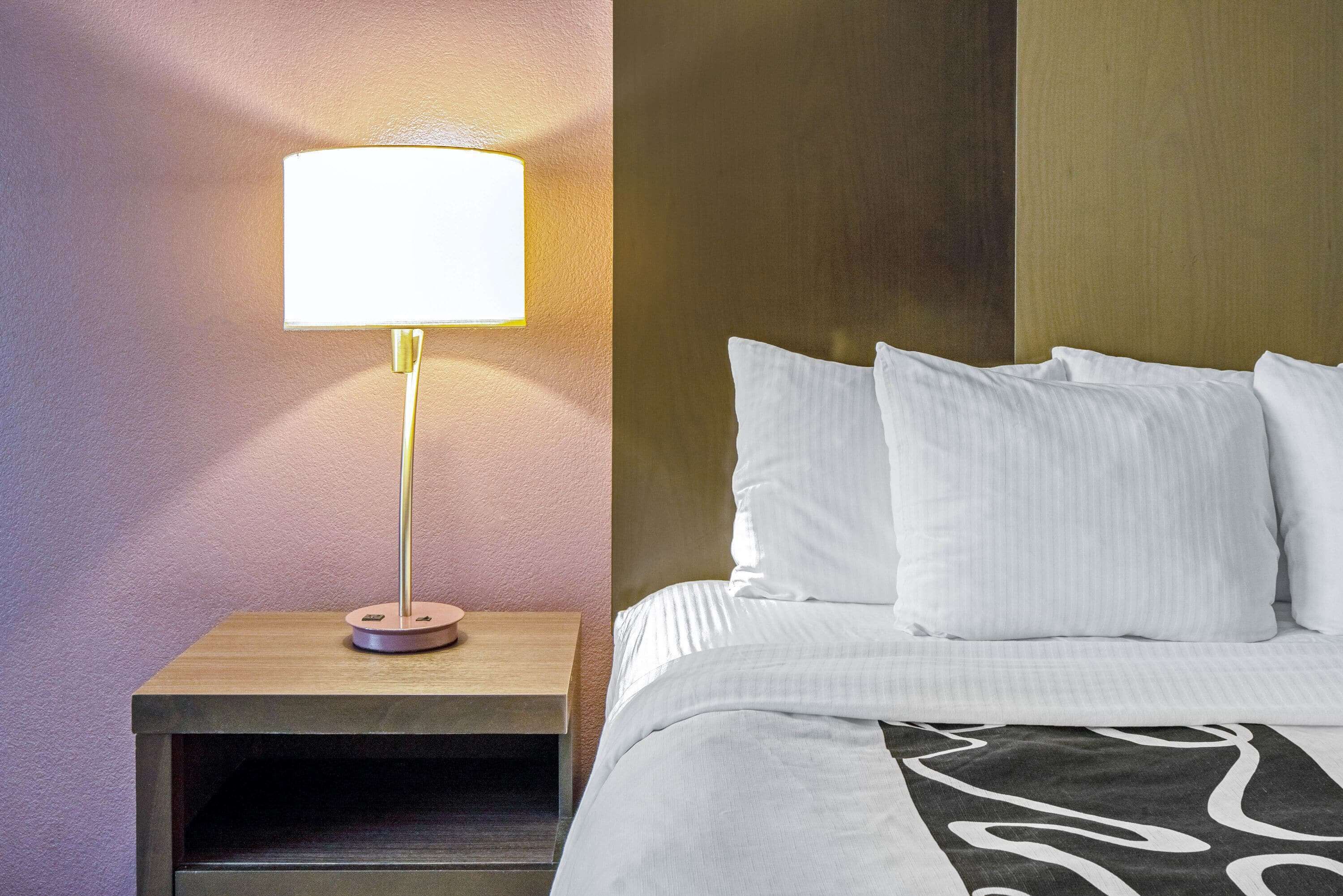 A neatly made bed with white linens and a black and white patterned throw. A bedside lamp with a white shade is on a wooden nightstand.