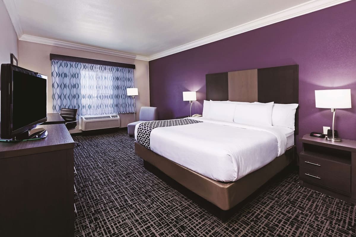 A hotel room with a large bed, a desk with a TV, a chair, a desk lamp, and a bedside table with a lamp.
