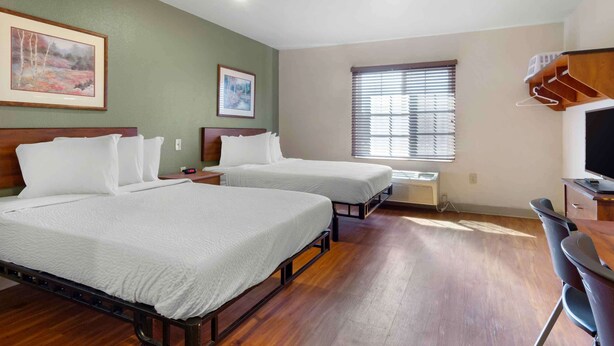 Quarto de hotel com duas camas, uma escrivaninha, uma cadeira, uma televisão e uma janela com persianas.